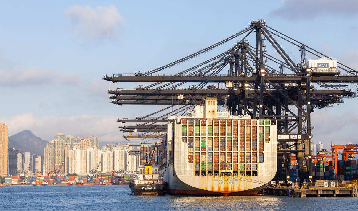 Container ship docked at a Hong Kong port with cranes and city skyline in the background, representing international logistics and the ability to ship from Hong Kong.