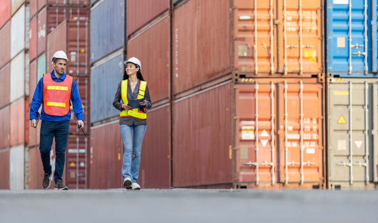 Two logistics professionals walk past shipping containers while discussing CBP enforcement and trade compliance strategy.