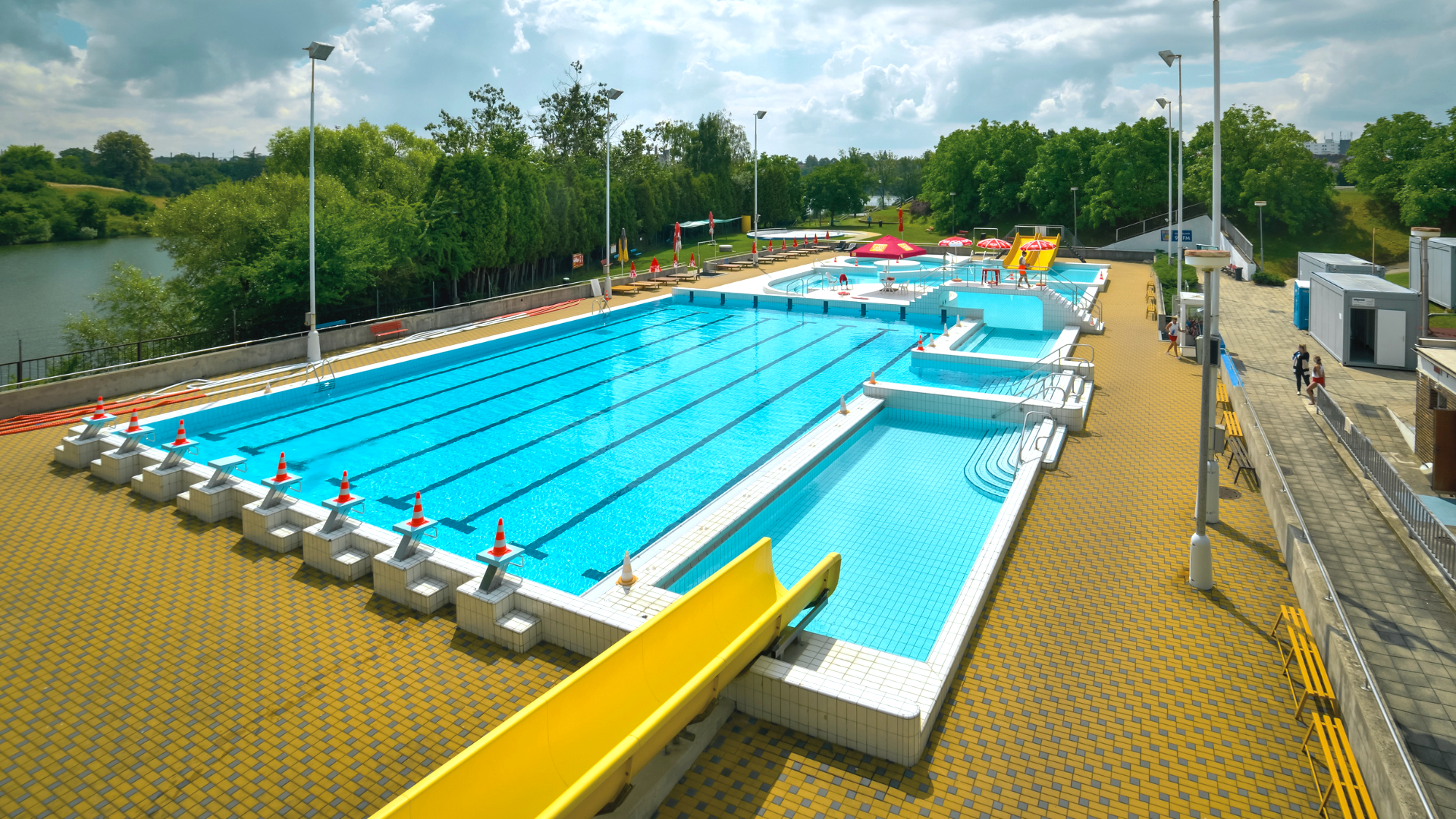 Outdoor swimming pool complex with several blue pools, yellow water slides, lane markers, lifeguard cones, and surrounding tiled deck, with trees, benches, and a cloudy sky in the background.