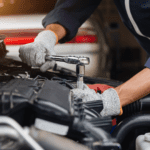 A mechanic wearing gloves uses a socket wrench to repair a car engine under the open hood in an auto workshop.