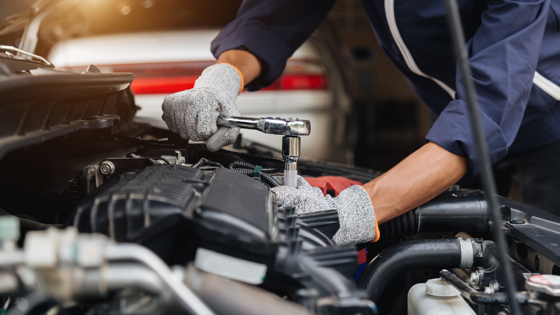 A mechanic wearing gloves uses a socket wrench to repair a car engine under the open hood in an auto workshop.