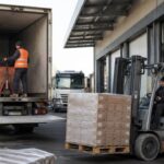 Forklift moving palletized cargo beside a truck at a warehouse loading area